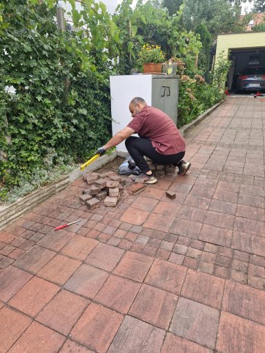 Pflege des Pflasterrands neben der Hecke Mann bearbeitet den Rand eines Pflasterwegs neben einer Hecke und einem hellen Versorgungskasten.