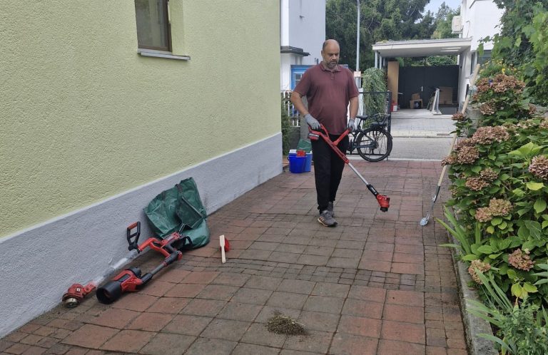 Gartenweg säubern Mann mit rotem Besen säubert einen gepflasterten Gartenweg; Gartengeräte liegen neben der Hauswand.