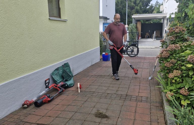 Pflege der Außenfläche Mann kehrt Pflastersteine vor einer grün gestrichenen Hauswand.