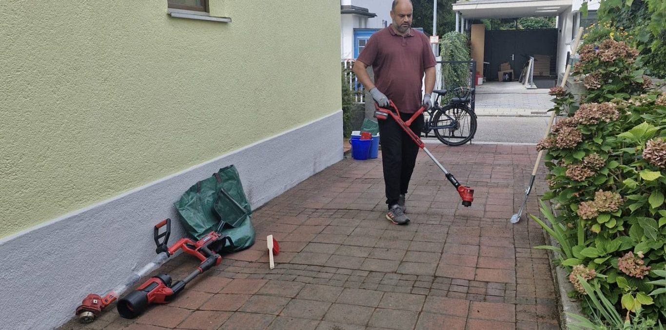 Gartenpflege am Haus Mitarbeiter mit Gartengerät auf einem gepflasterten Weg neben einer Hauswand.