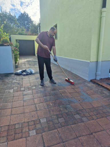 Pflasterreinigung mit dem Besen Mann mit rotem Besen kehrt eine gepflasterte Fläche vor einer grün gestrichenen Hauswand.