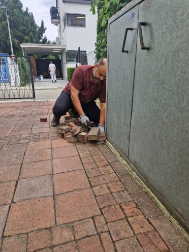 Arbeiten am Pflasterrand an der Hauswand Mann kniet an einer Hauswand und nimmt Pflastersteine am Rand eines gepflasterten Wegs auf.