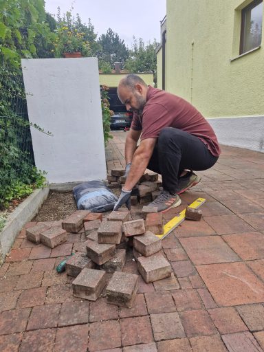 Detailaufnahme am Pflasterrand Mann in rotem Shirt kniet neben einem Versorgungskasten und arbeitet an aufgenommenen Pflastersteinen.